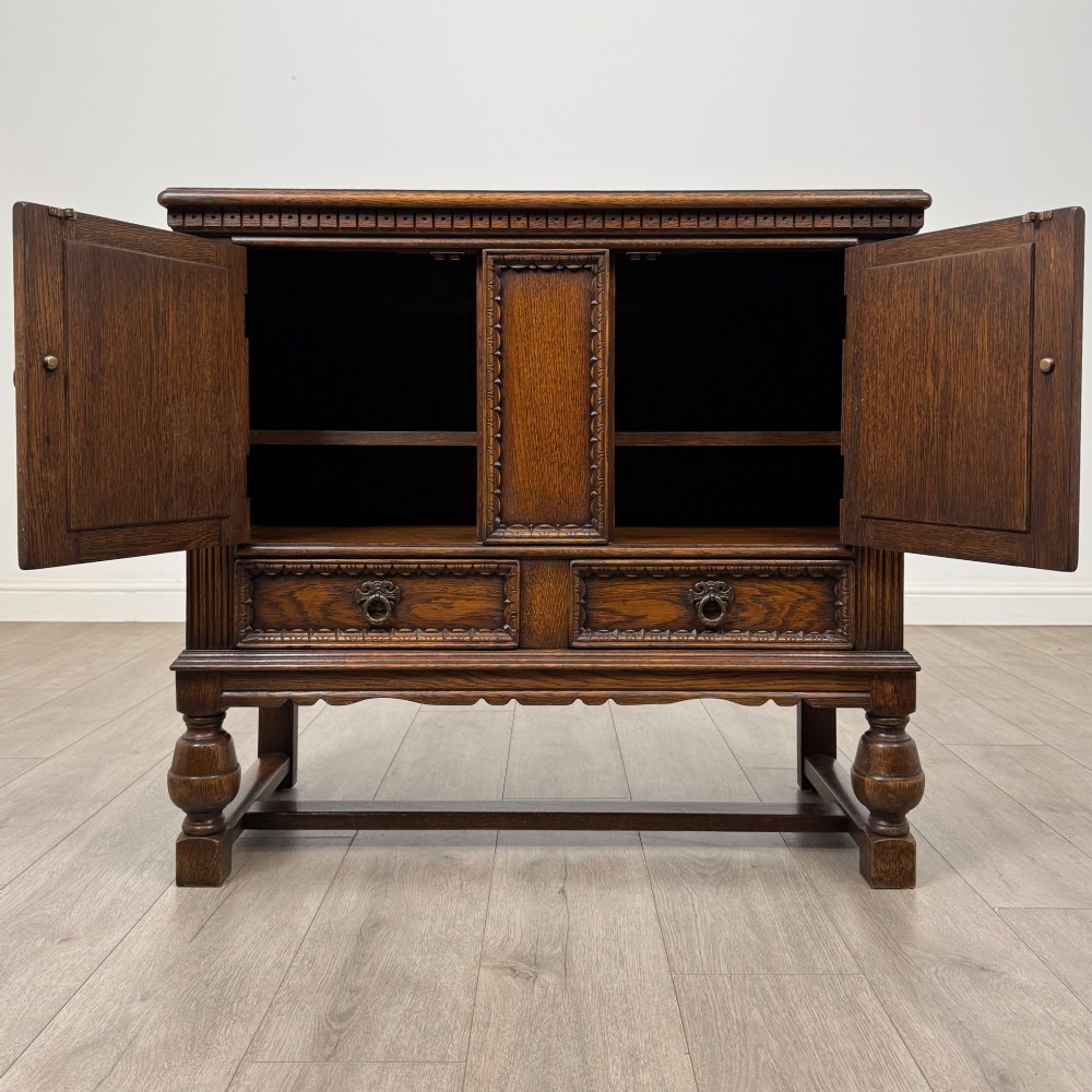 A dark oak cupboard featuring two relief-carved heraldic door panels above two drawers, supported by turned legs and stretchers. View II