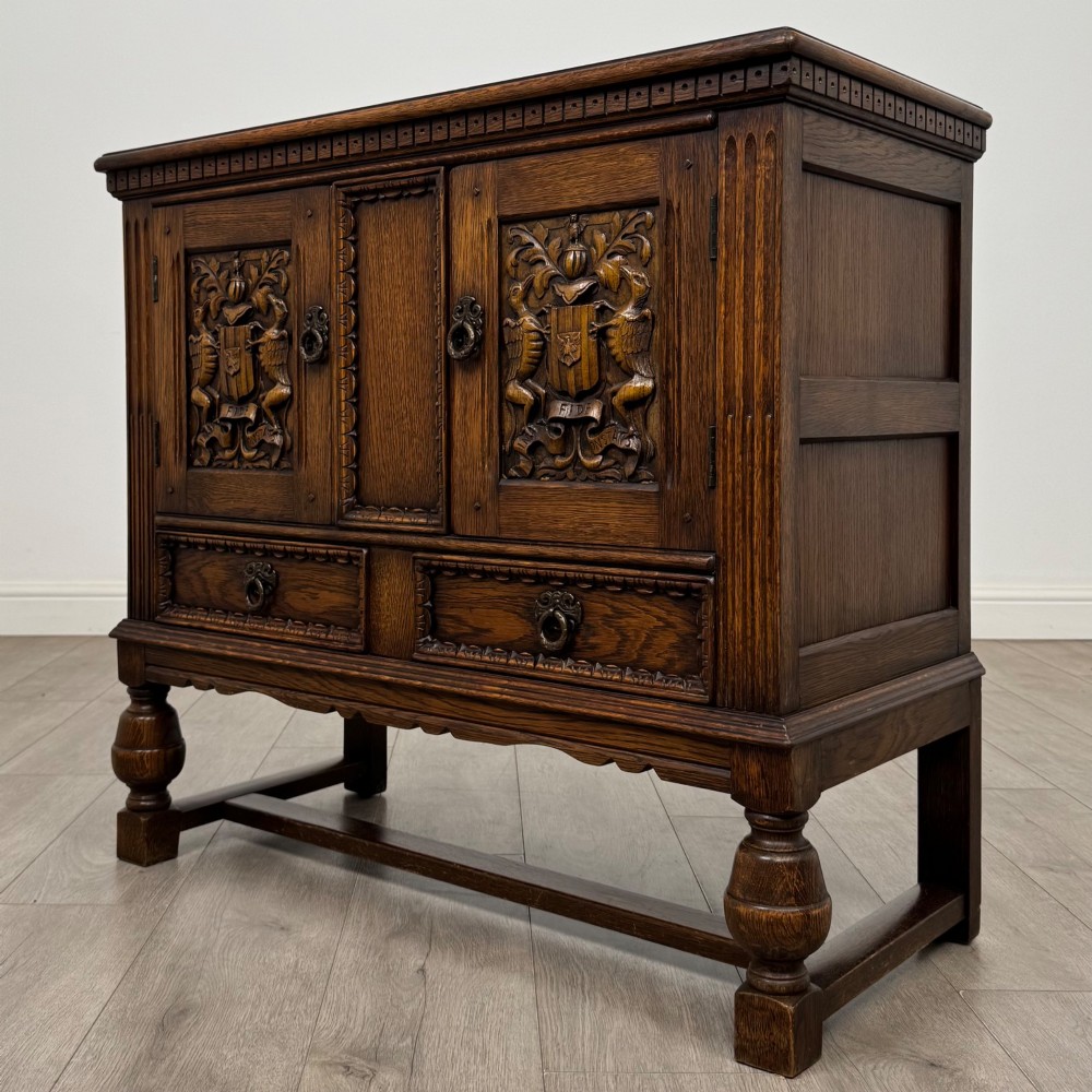 A dark oak cupboard featuring two relief-carved heraldic door panels above two drawers, supported by turned legs and stretchers. View III