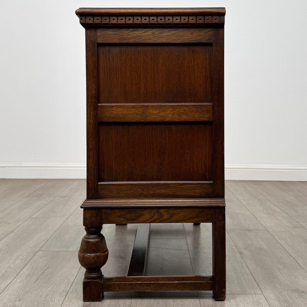 A dark oak cupboard featuring two relief-carved heraldic door panels above two drawers, supported by turned legs and stretchers. View IV