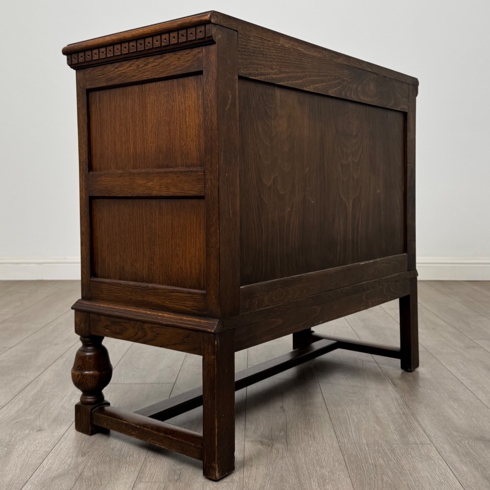 A dark oak cupboard featuring two relief-carved heraldic door panels above two drawers, supported by turned legs and stretchers. View V