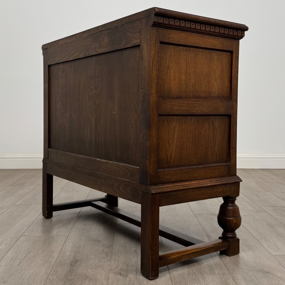A dark oak cupboard featuring two relief-carved heraldic door panels above two drawers, supported by turned legs and stretchers. View VII