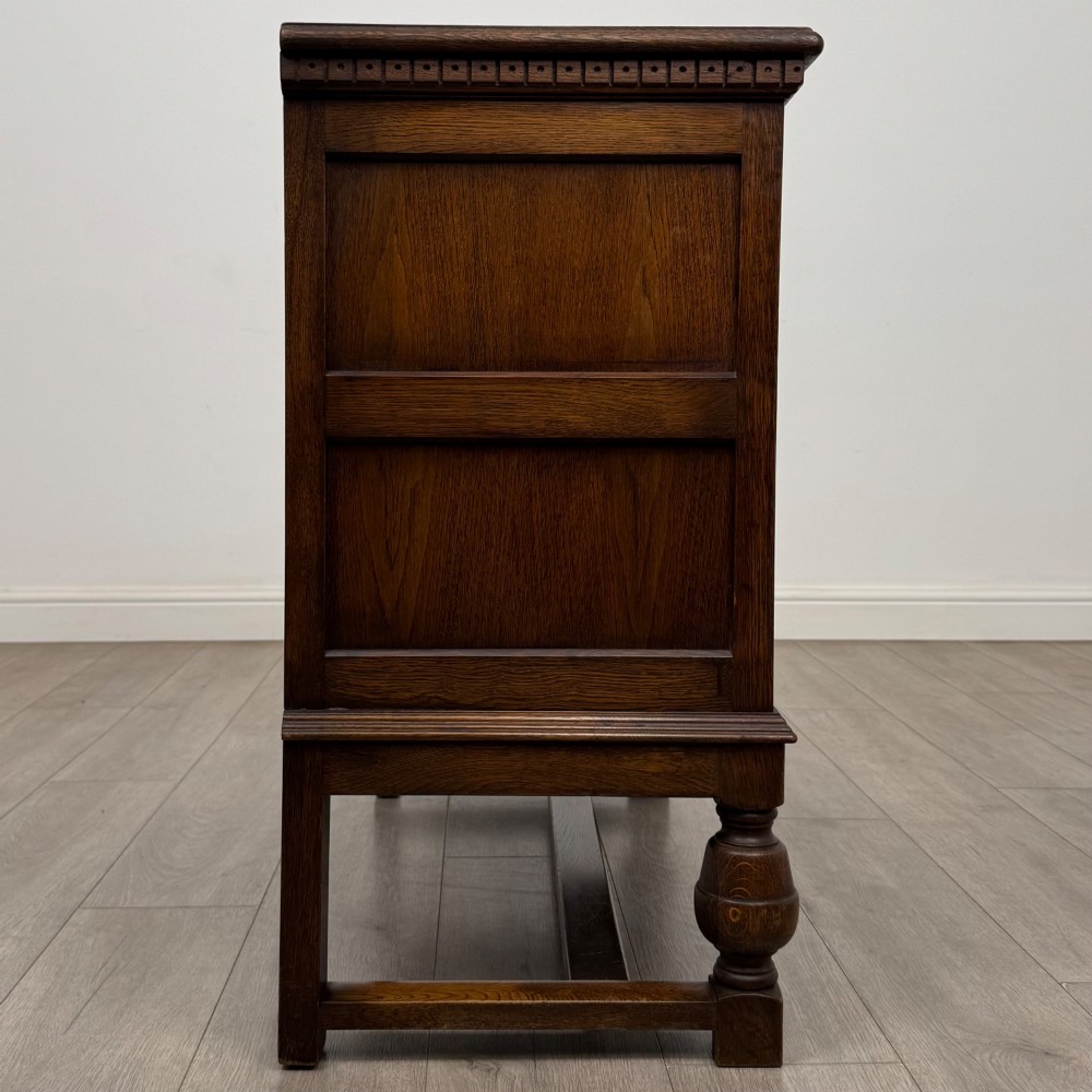 A dark oak cupboard featuring two relief-carved heraldic door panels above two drawers, supported by turned legs and stretchers. View VIII