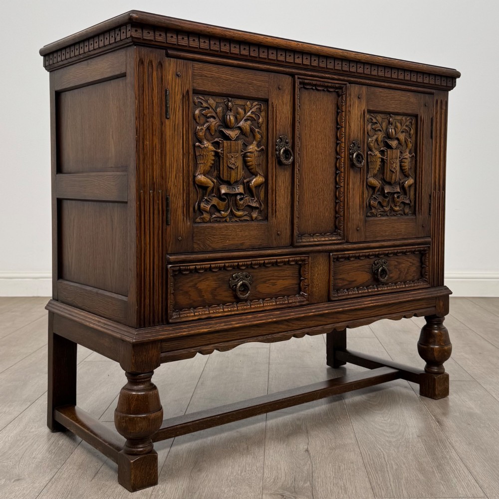 A dark oak cupboard featuring two relief-carved heraldic door panels above two drawers, supported by turned legs and stretchers. View IX