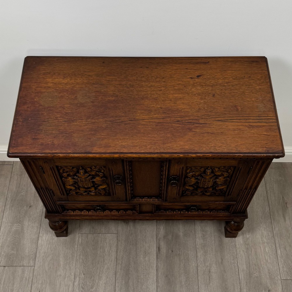 A dark oak cupboard featuring two relief-carved heraldic door panels above two drawers, supported by turned legs and stretchers. View X