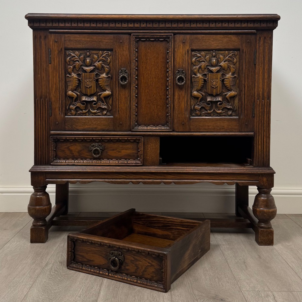 A dark oak cupboard featuring two relief-carved heraldic door panels above two drawers, supported by turned legs and stretchers. View XI