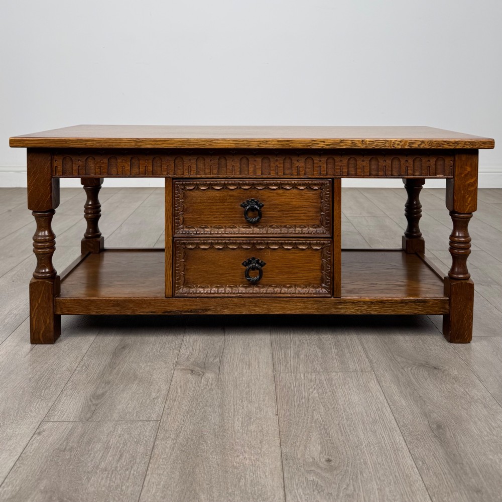 A rectangular carved oak coffee table with two central drawers, metal drop handles, turned legs, and a lower shelf.