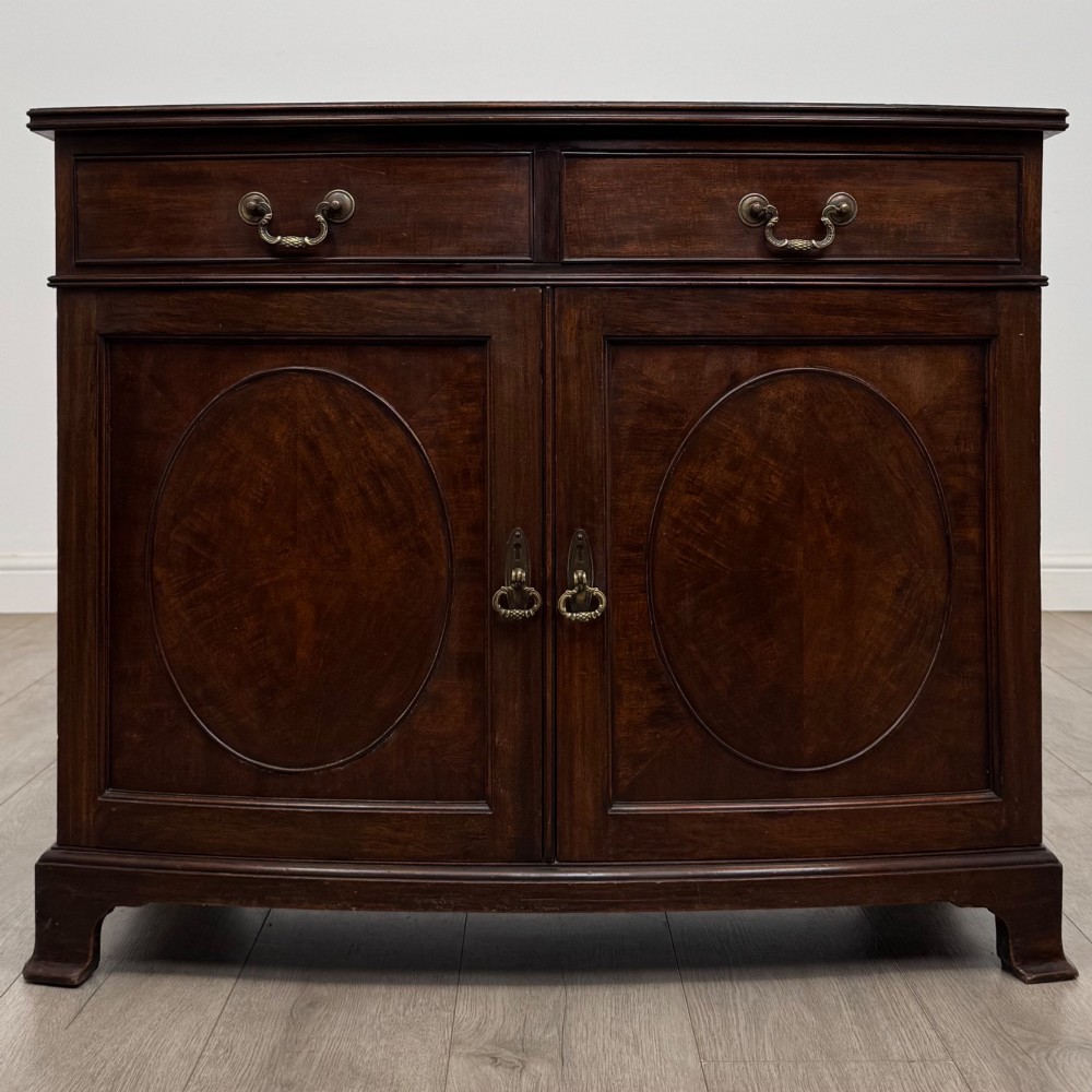 An antique Edwardian mahogany bow-front cupboard featuring two upper drawers with brass handles and two lower doors with oval wood grain panels.