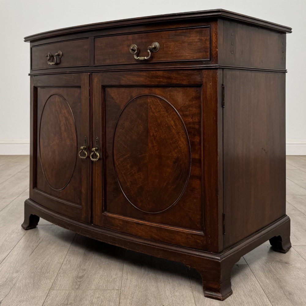 An antique Edwardian mahogany bow-front cupboard featuring two upper drawers with brass handles and two lower doors with oval wood grain panels. View III