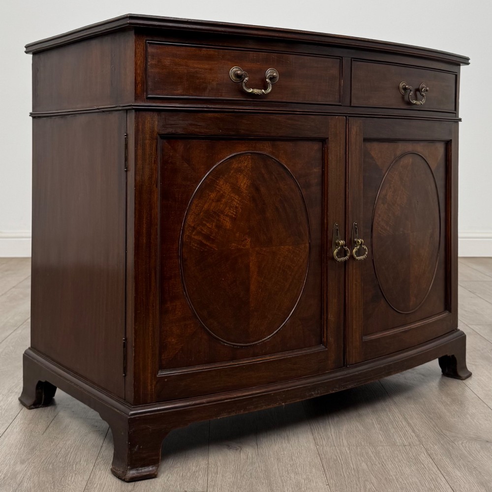 An antique Edwardian mahogany bow-front cupboard featuring two upper drawers with brass handles and two lower doors with oval wood grain panels. View IX