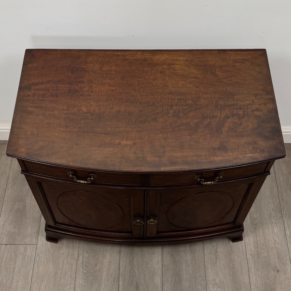 An antique Edwardian mahogany bow-front cupboard featuring two upper drawers with brass handles and two lower doors with oval wood grain panels. View X