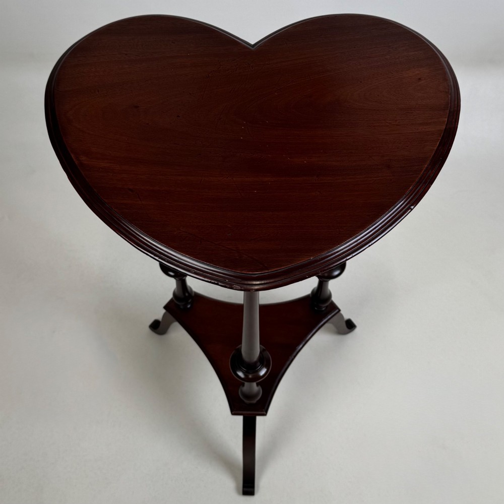 A tall dark mahogany side table featuring a heart-shaped top with a raised rim, three slender turned spindle legs, a lower triangular shelf, and flared feet. View V