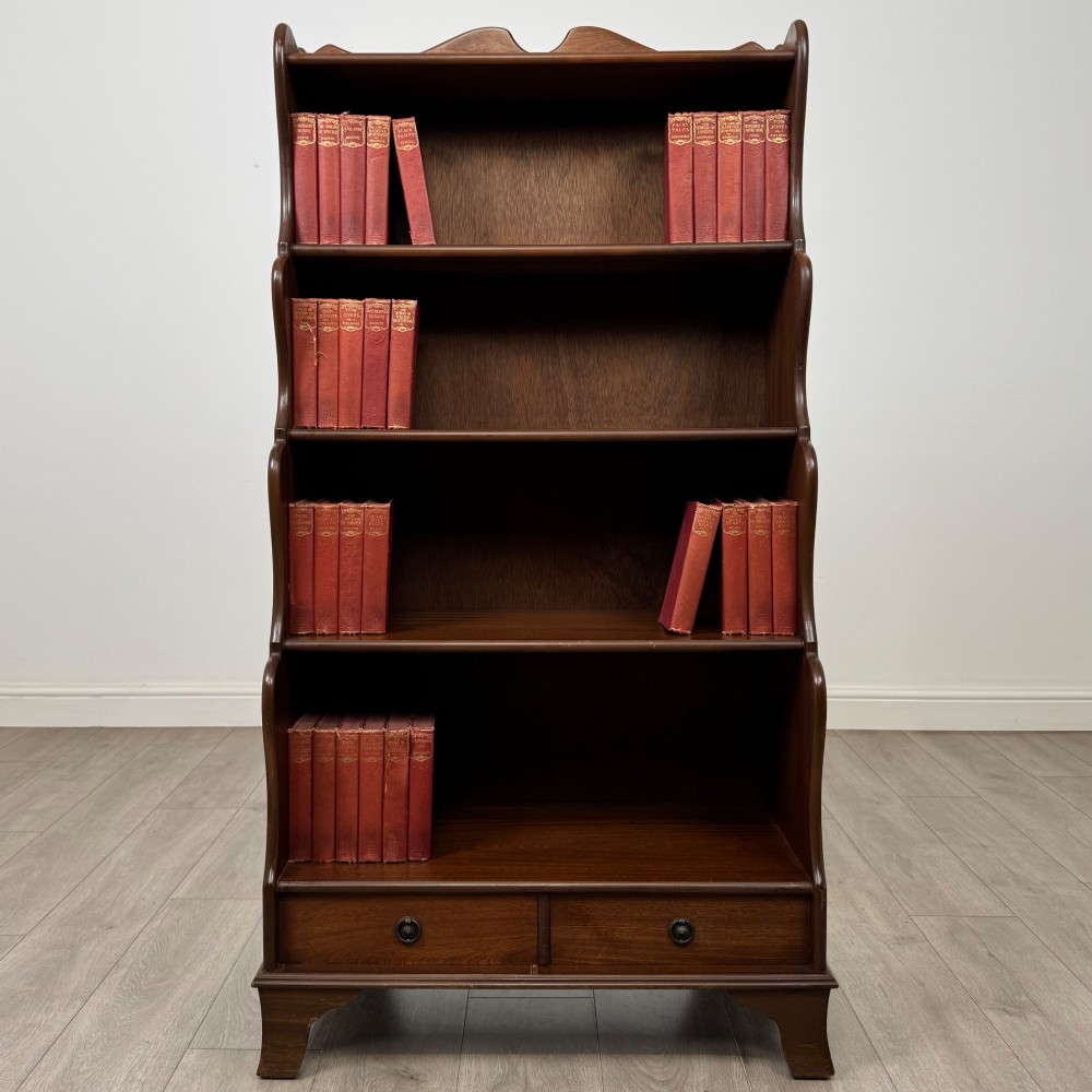 A freestanding dark mahogany waterfall bookcase with four graduated open shelves above two base drawers featuring ring pull handles.