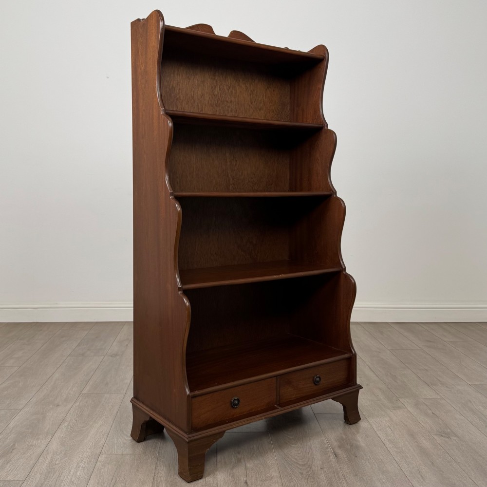 A freestanding dark mahogany waterfall bookcase with four graduated open shelves above two base drawers featuring ring pull handles. View IX