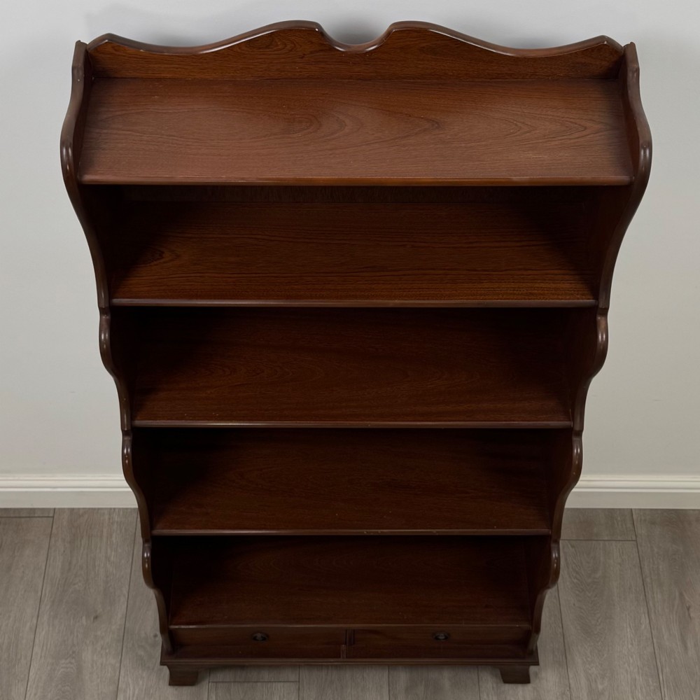 A freestanding dark mahogany waterfall bookcase with four graduated open shelves above two base drawers featuring ring pull handles. View X
