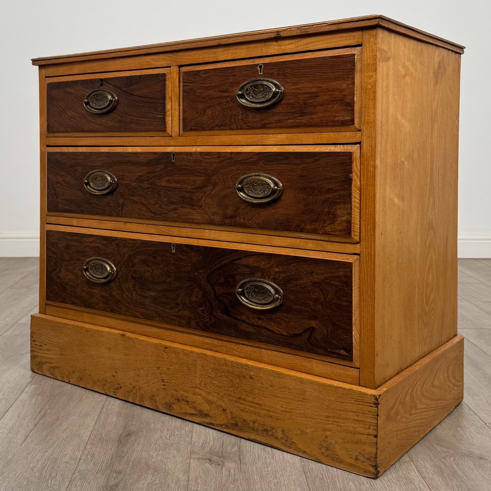 An antique Victorian oak chest of drawers featuring two short top drawers over two long lower drawers with dark drawer fronts, brass oval handles, and a plinth base. View II