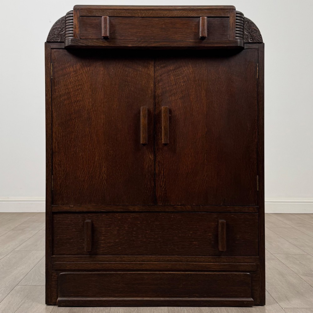 A dark oak Art Deco storage cupboard featuring a stepped top structure, carved rounded shoulders, double doors, and timber bar handles.