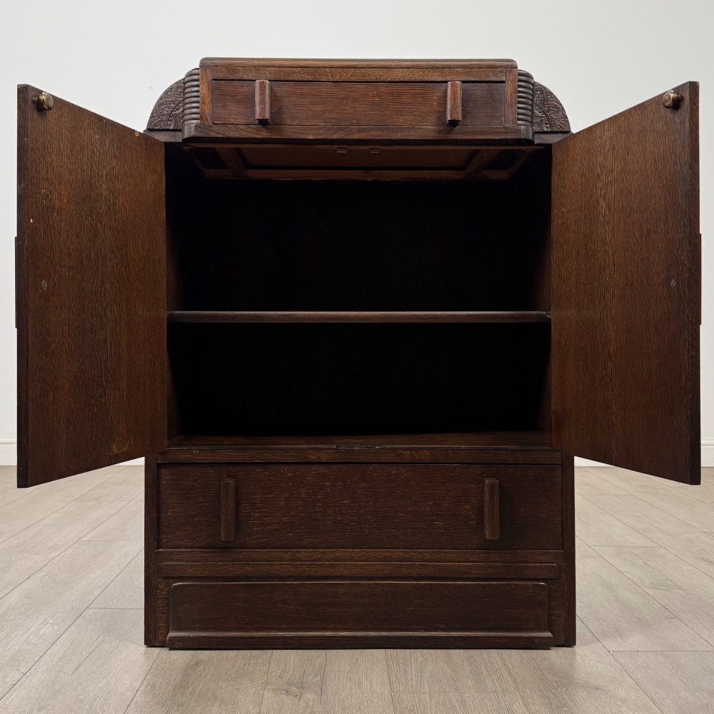 A dark oak Art Deco storage cupboard featuring a stepped top structure, carved rounded shoulders, double doors, and timber bar handles. View II