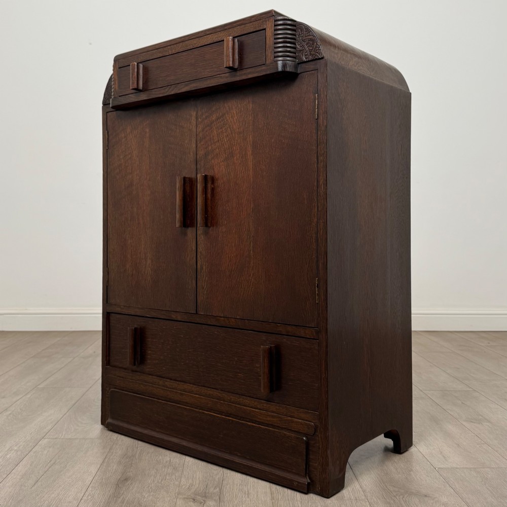 A dark oak Art Deco storage cupboard featuring a stepped top structure, carved rounded shoulders, double doors, and timber bar handles. View III