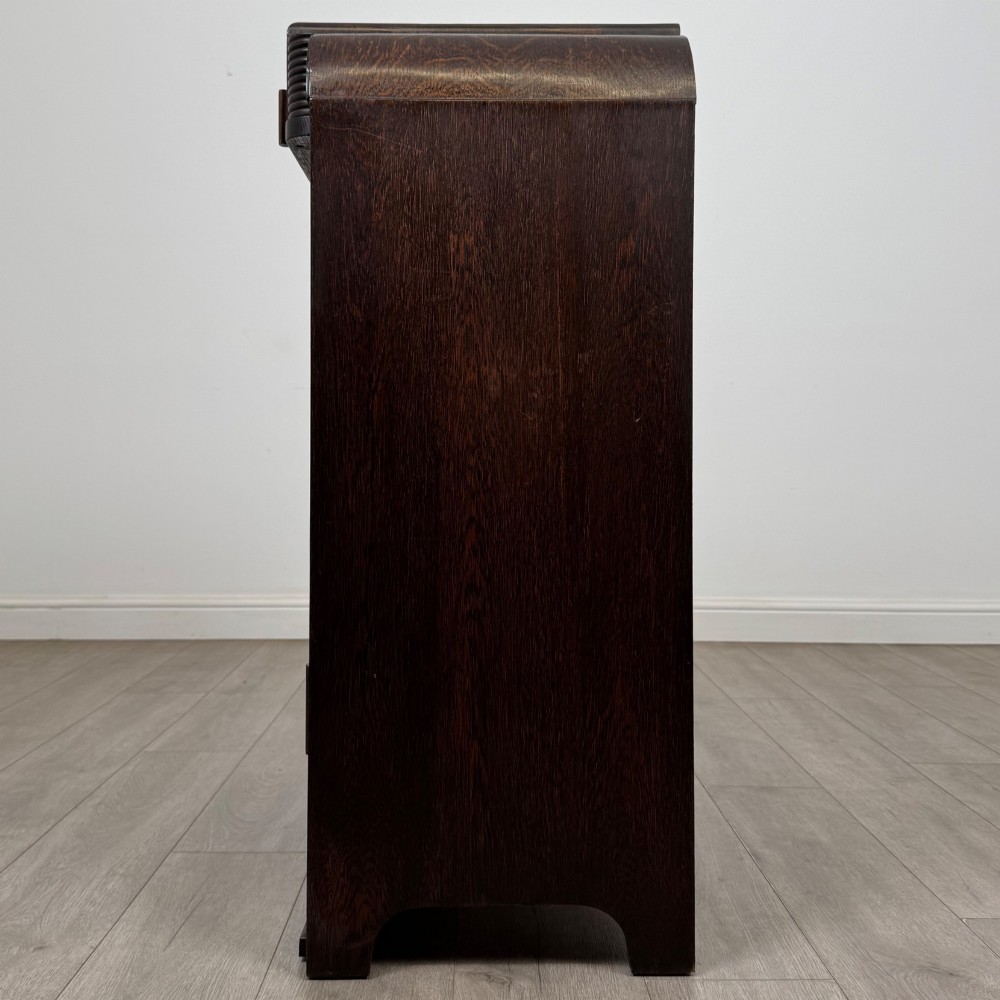 A dark oak Art Deco storage cupboard featuring a stepped top structure, carved rounded shoulders, double doors, and timber bar handles. View IV