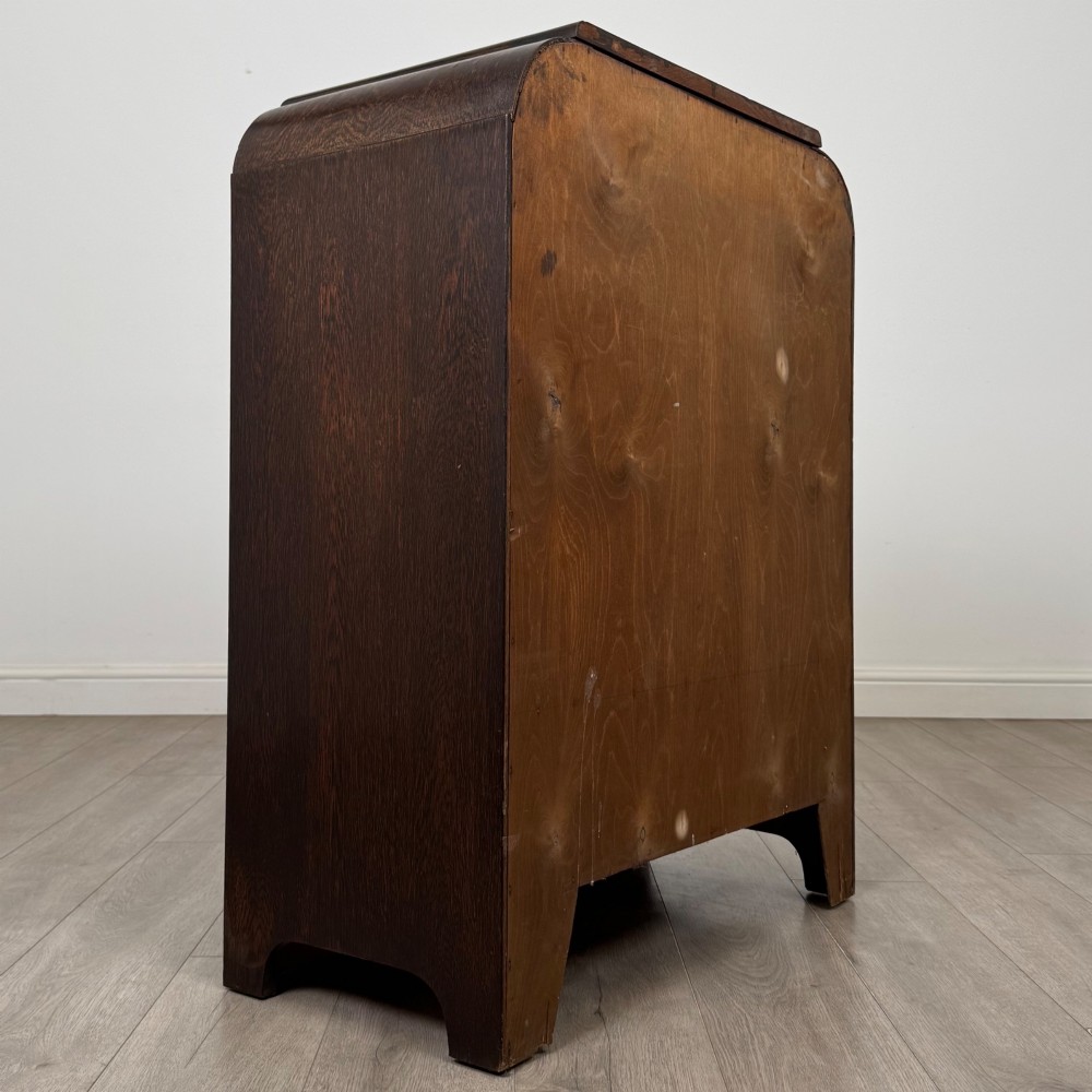 A dark oak Art Deco storage cupboard featuring a stepped top structure, carved rounded shoulders, double doors, and timber bar handles. View V