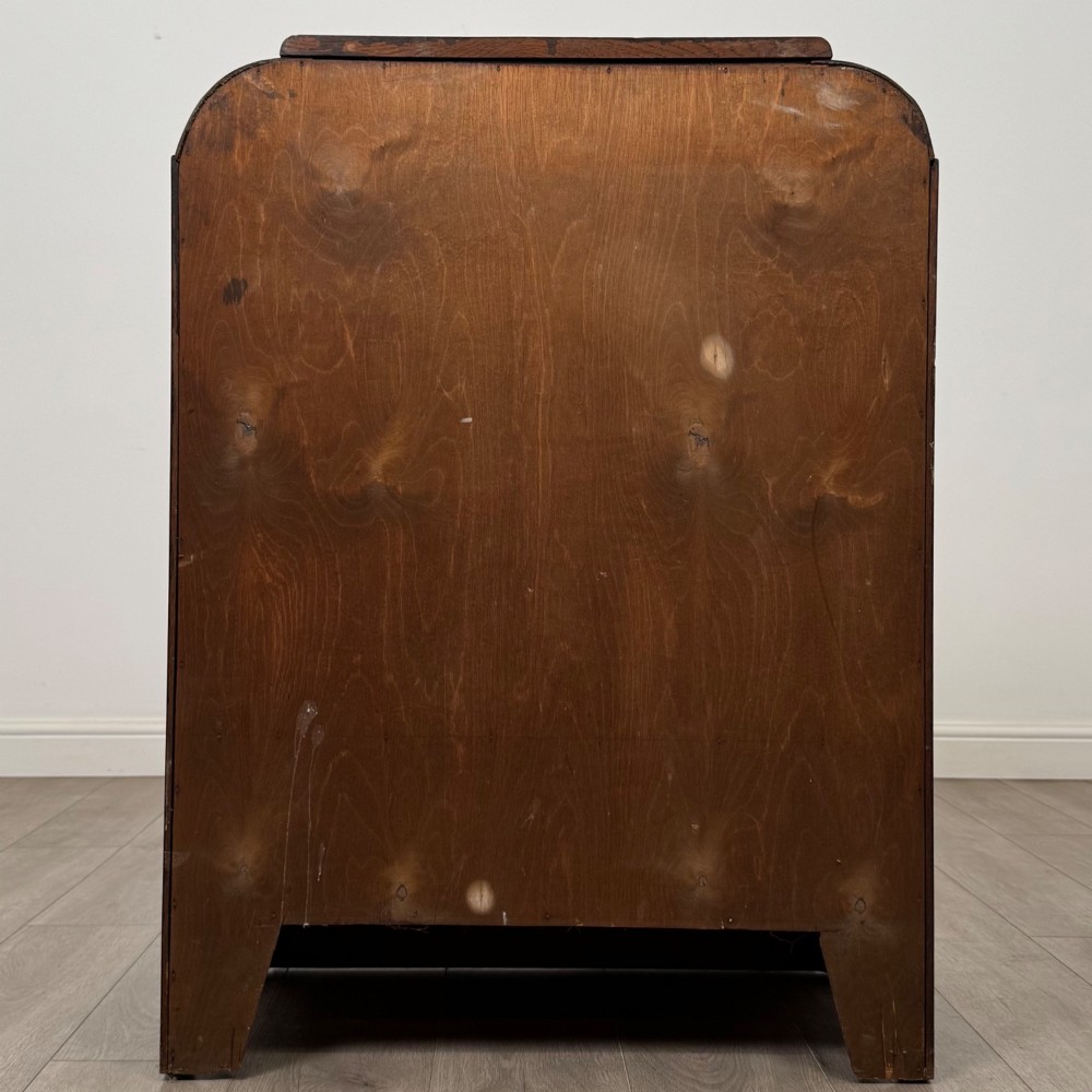 A dark oak Art Deco storage cupboard featuring a stepped top structure, carved rounded shoulders, double doors, and timber bar handles. View VI