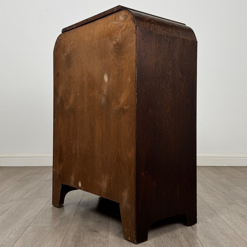 A dark oak Art Deco storage cupboard featuring a stepped top structure, carved rounded shoulders, double doors, and timber bar handles. View VII