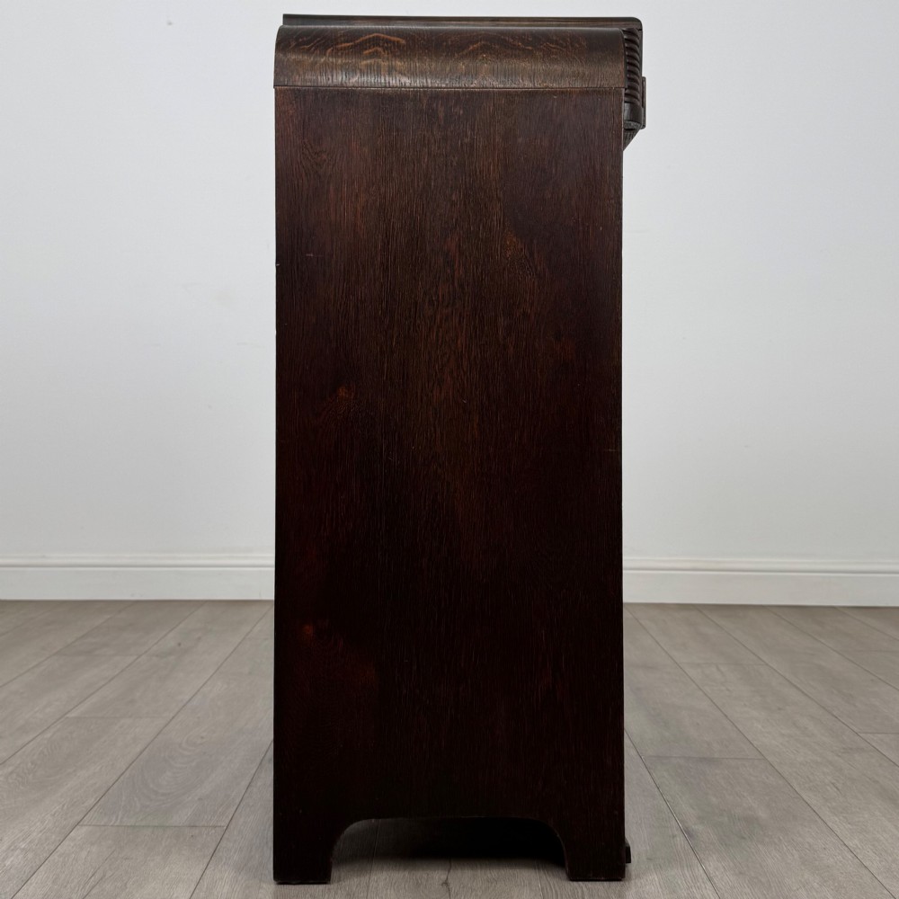 A dark oak Art Deco storage cupboard featuring a stepped top structure, carved rounded shoulders, double doors, and timber bar handles. View VIII