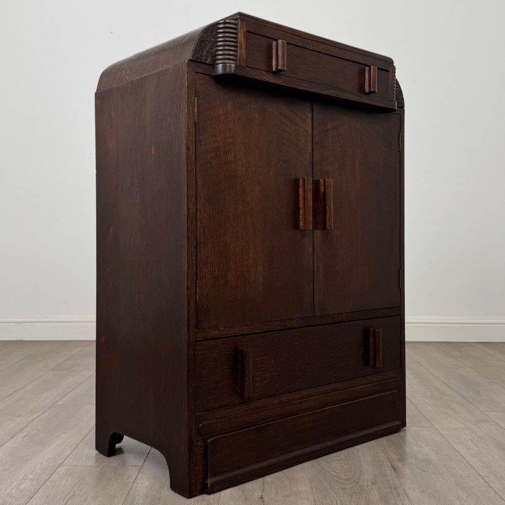 A dark oak Art Deco storage cupboard featuring a stepped top structure, carved rounded shoulders, double doors, and timber bar handles. View IX