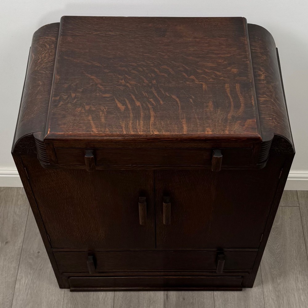 A dark oak Art Deco storage cupboard featuring a stepped top structure, carved rounded shoulders, double doors, and timber bar handles. View X
