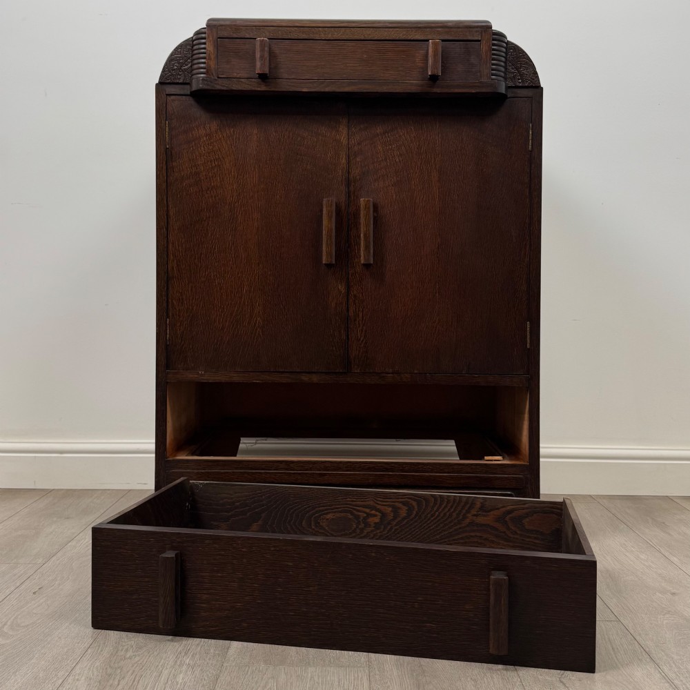 A dark oak Art Deco storage cupboard featuring a stepped top structure, carved rounded shoulders, double doors, and timber bar handles. View XI