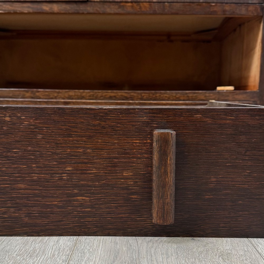 A dark oak Art Deco storage cupboard featuring a stepped top structure, carved rounded shoulders, double doors, and timber bar handles. View XII