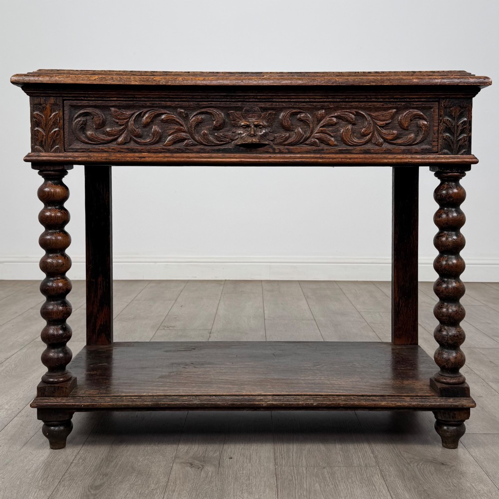 A dark oak Victorian side table featuring bobbin-turned legs, a lower shelf, and a carved frieze with a central mask motif.
