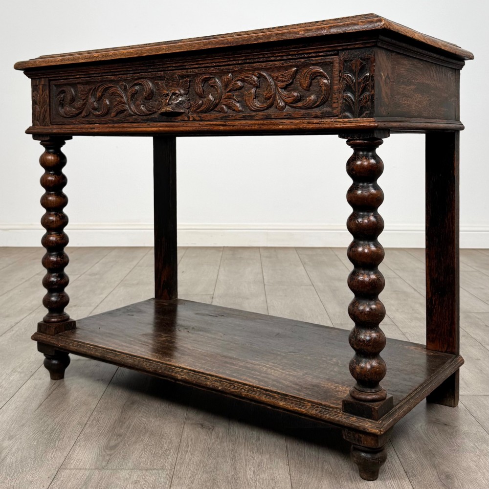 A dark oak Victorian side table featuring bobbin-turned legs, a lower shelf, and a carved frieze with a central mask motif. View II