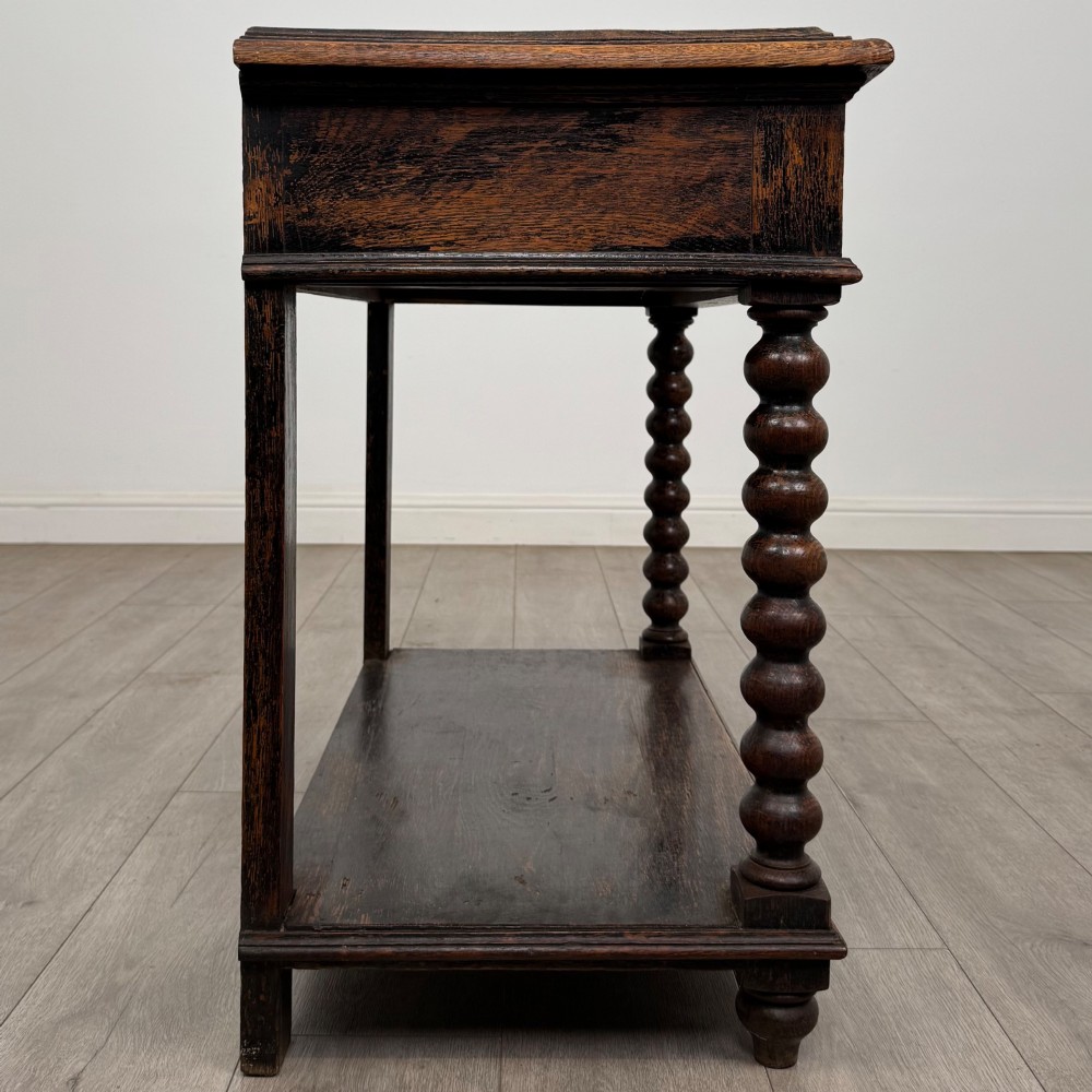 A dark oak Victorian side table featuring bobbin-turned legs, a lower shelf, and a carved frieze with a central mask motif. View VII