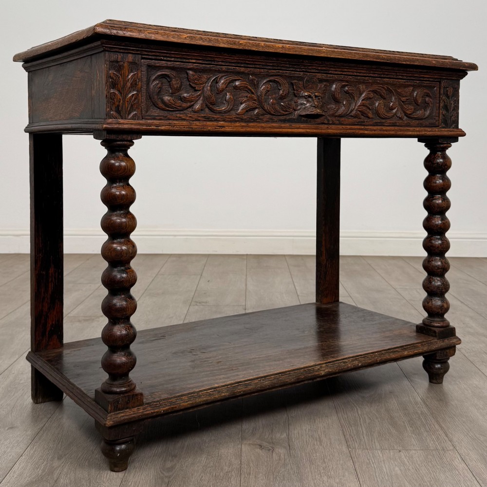 A dark oak Victorian side table featuring bobbin-turned legs, a lower shelf, and a carved frieze with a central mask motif. View VIII