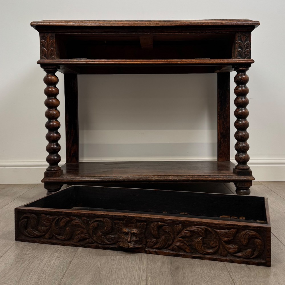 A dark oak Victorian side table featuring bobbin-turned legs, a lower shelf, and a carved frieze with a central mask motif. View X