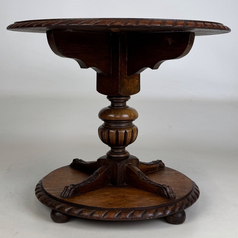 A round dark oak coffee table featuring a carved edge, a turned central pedestal, and a circular base supported on bun feet.