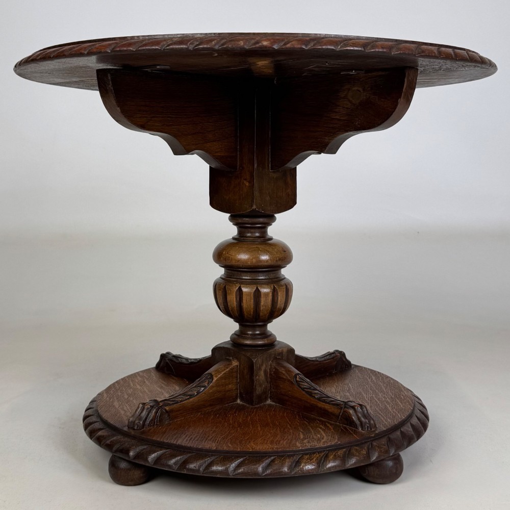 A round dark oak coffee table featuring a carved edge, a turned central pedestal, and a circular base supported on bun feet. View III