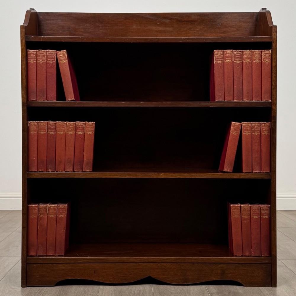 A dark mahogany open bookcase featuring three broad fixed shelves, shaped gallery side panels, and a subtle wave-formed lower apron.