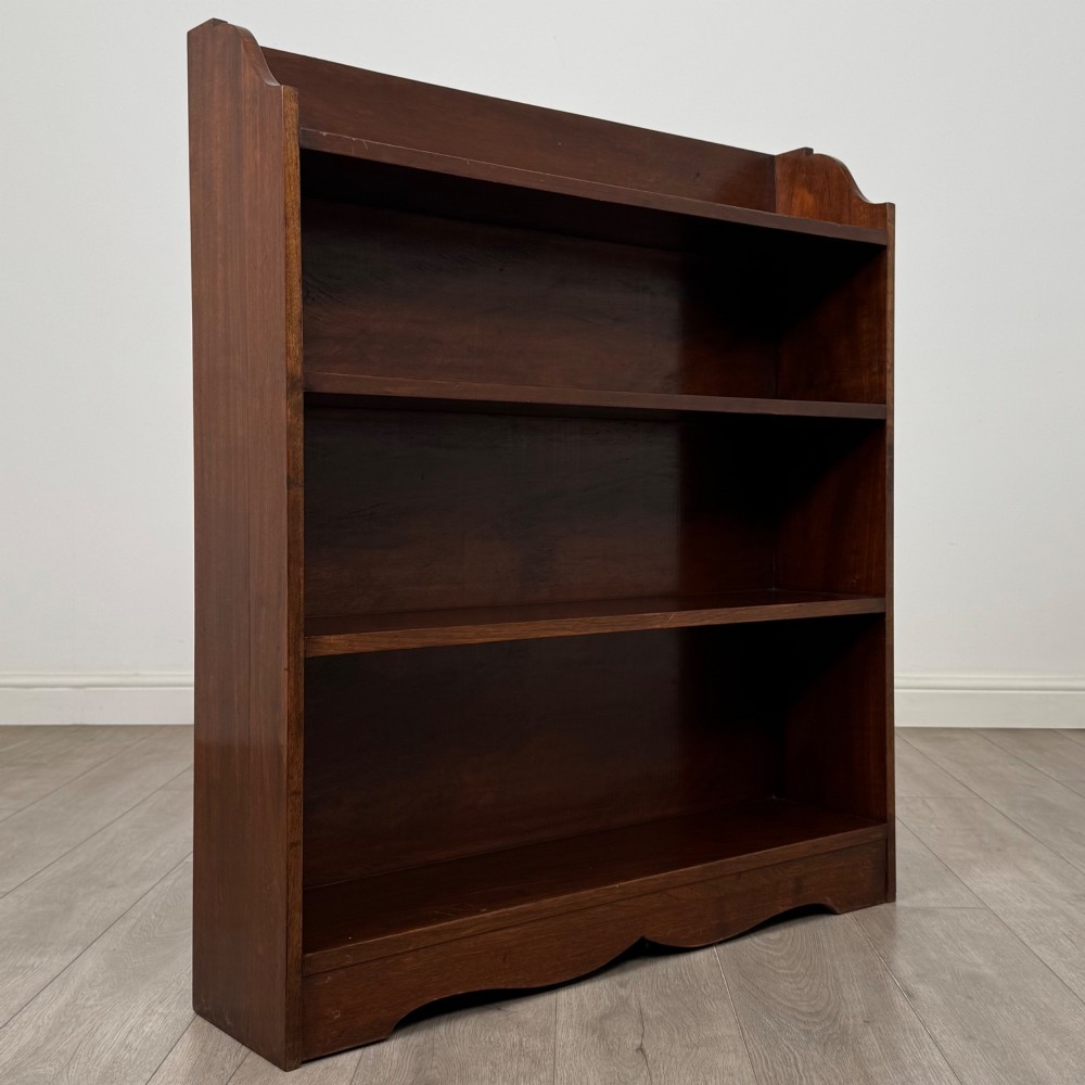A dark mahogany open bookcase featuring three broad fixed shelves, shaped gallery side panels, and a subtle wave-formed lower apron. View IX