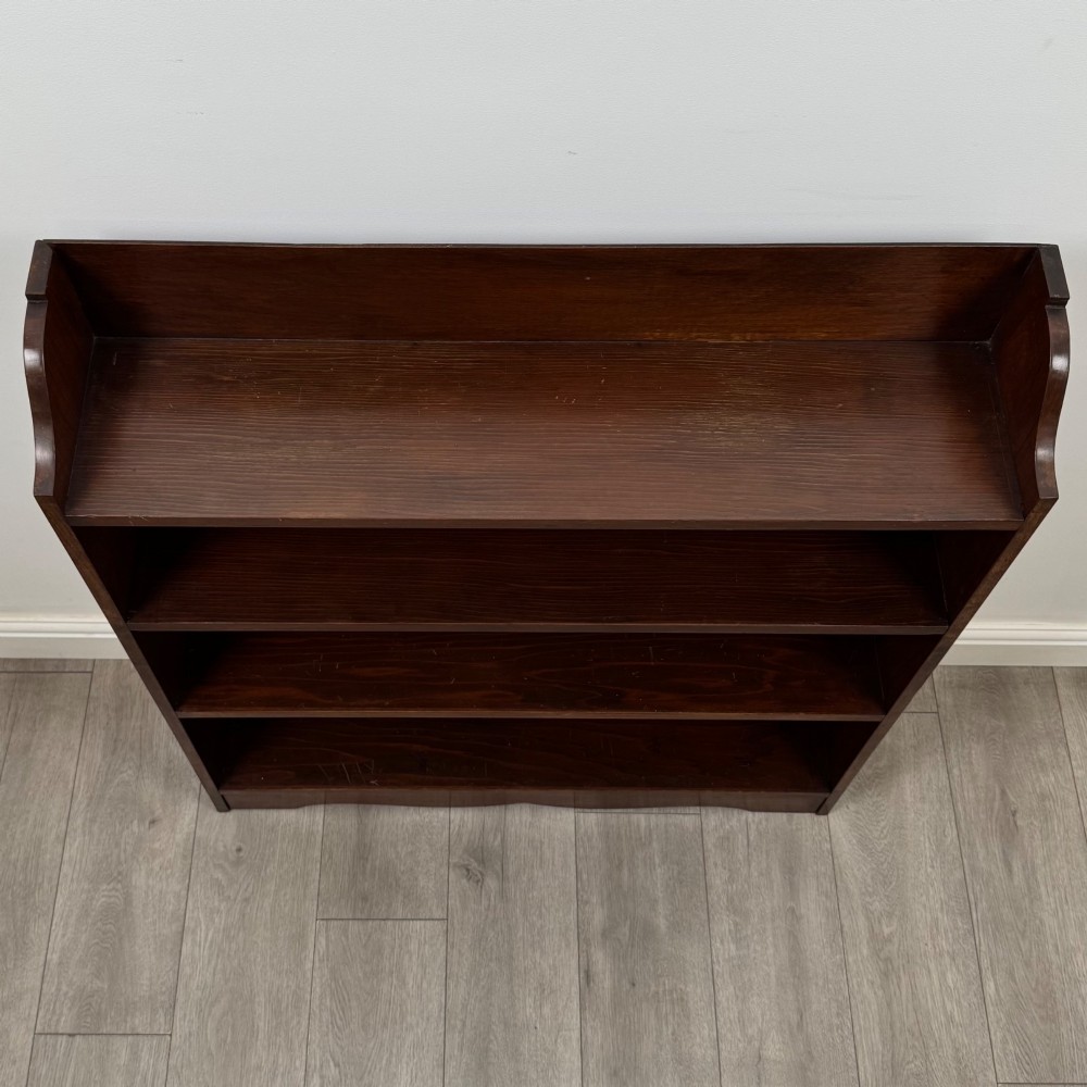 A dark mahogany open bookcase featuring three broad fixed shelves, shaped gallery side panels, and a subtle wave-formed lower apron. View X