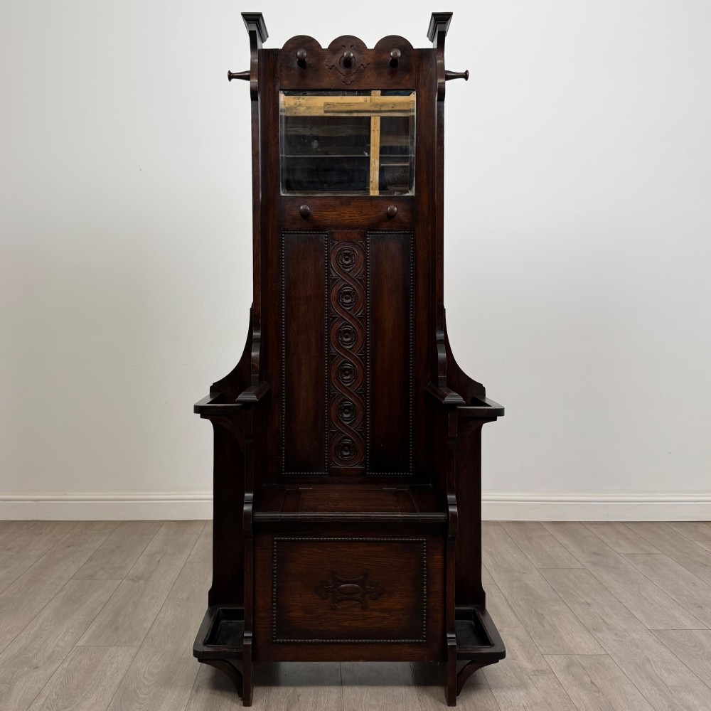 A tall dark oak Edwardian hall stand featuring carved panels, turned coat pegs, a central mirror, a lift-top storage seat, and umbrella holders on both sides.