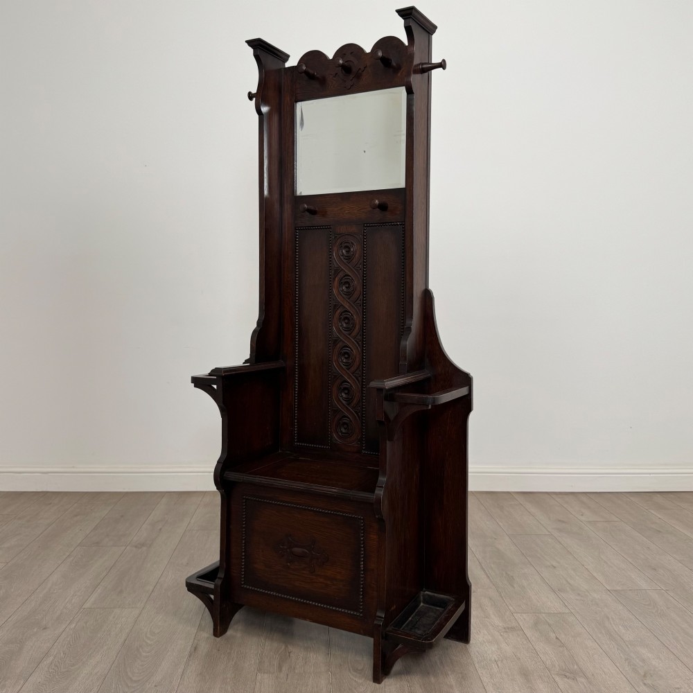 A tall dark oak Edwardian hall stand featuring carved panels, turned coat pegs, a central mirror, a lift-top storage seat, and umbrella holders on both sides. View II