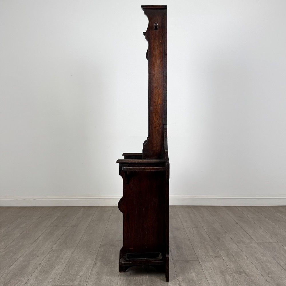 A tall dark oak Edwardian hall stand featuring carved panels, turned coat pegs, a central mirror, a lift-top storage seat, and umbrella holders on both sides. View III