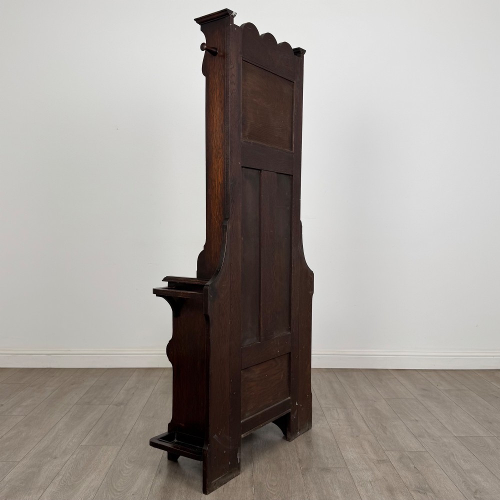 A tall dark oak Edwardian hall stand featuring carved panels, turned coat pegs, a central mirror, a lift-top storage seat, and umbrella holders on both sides. View IV