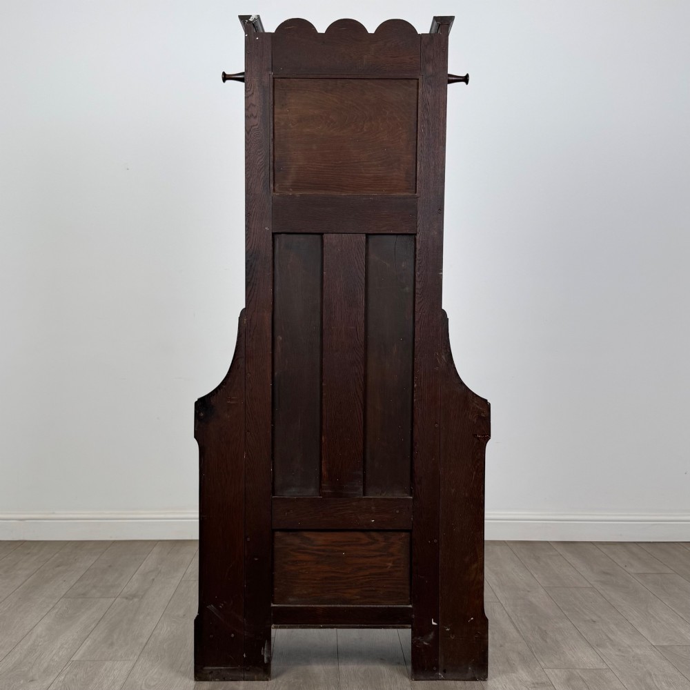 A tall dark oak Edwardian hall stand featuring carved panels, turned coat pegs, a central mirror, a lift-top storage seat, and umbrella holders on both sides. View V
