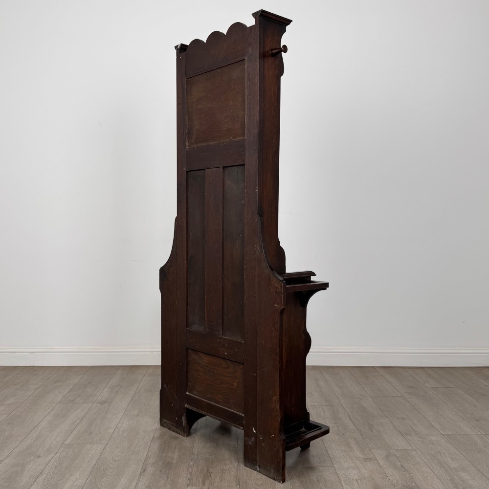 A tall dark oak Edwardian hall stand featuring carved panels, turned coat pegs, a central mirror, a lift-top storage seat, and umbrella holders on both sides. View VI