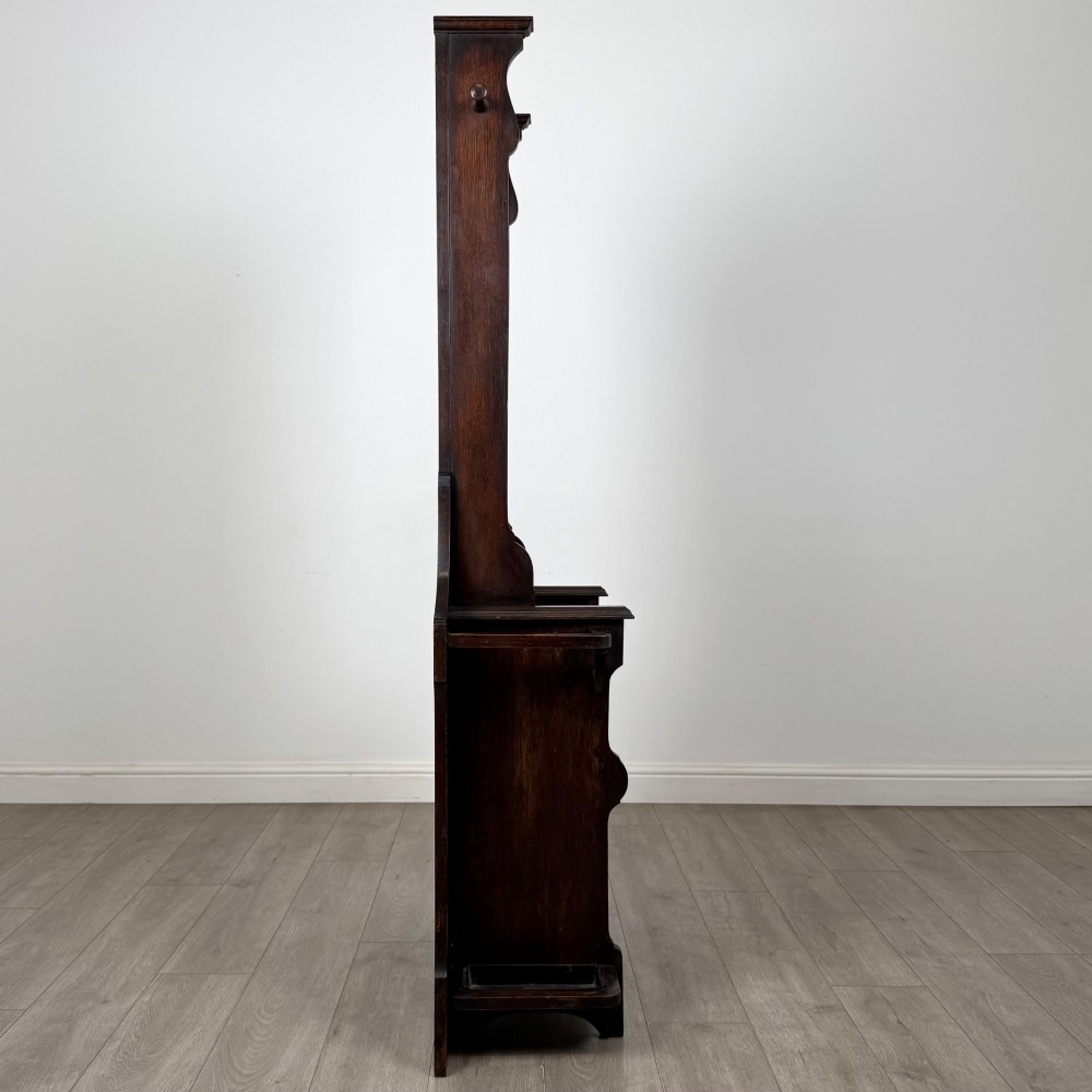 A tall dark oak Edwardian hall stand featuring carved panels, turned coat pegs, a central mirror, a lift-top storage seat, and umbrella holders on both sides. View VII
