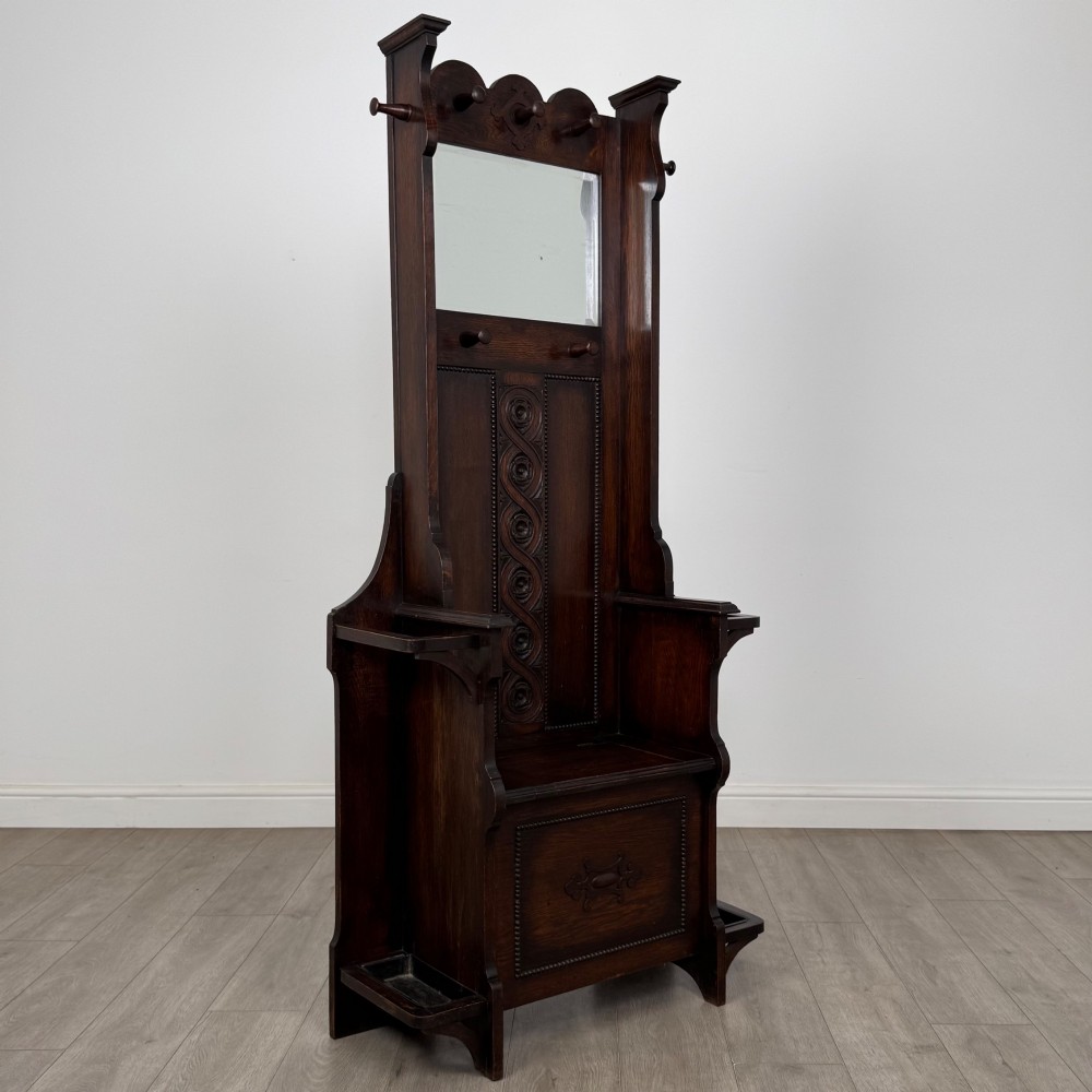 A tall dark oak Edwardian hall stand featuring carved panels, turned coat pegs, a central mirror, a lift-top storage seat, and umbrella holders on both sides. View VIII
