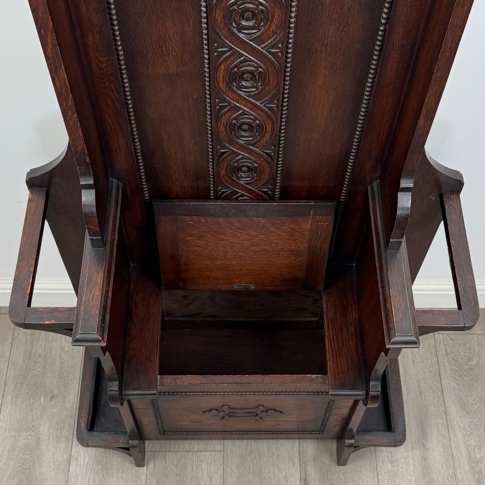 A tall dark oak Edwardian hall stand featuring carved panels, turned coat pegs, a central mirror, a lift-top storage seat, and umbrella holders on both sides. View X